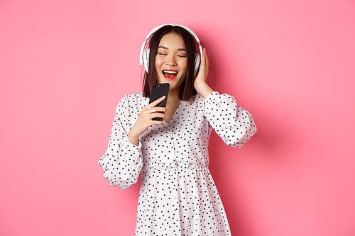 Cute asian woman playing karaoke app, singing in mobile phone and using headphones, standing in dress over pink background.