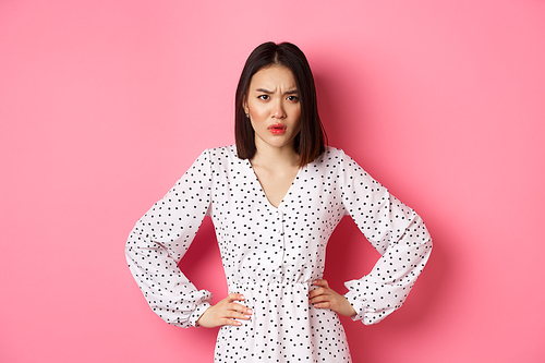 Angry asian woman staring at camera frustrated, holding hands on waist and waiting for explanations, standing in dress against pink background.