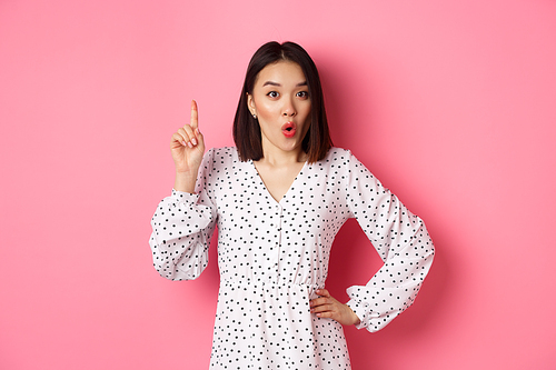 Excited asian girl having an idea, saying suggestion, raising finger and talking, standing over pink background.