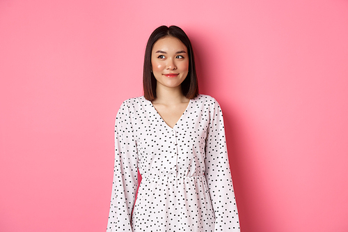 Cute asian woman smiling, looking left at copy space, standing on romantic pink background.