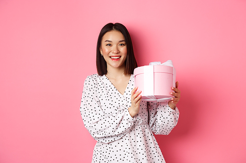 Romantic asian woman in cute dress holding box with gift, smiling happy at camera, standing with present over pink background.