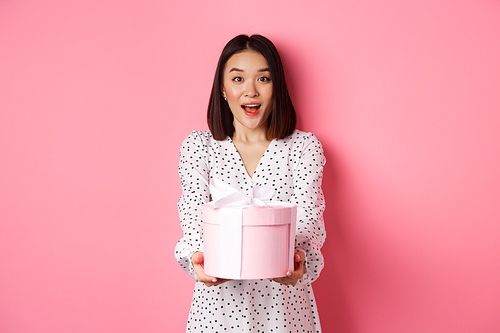 Cute asian woman congratulate with holiday or birthday, giving gift in cute box, standing over pink background.