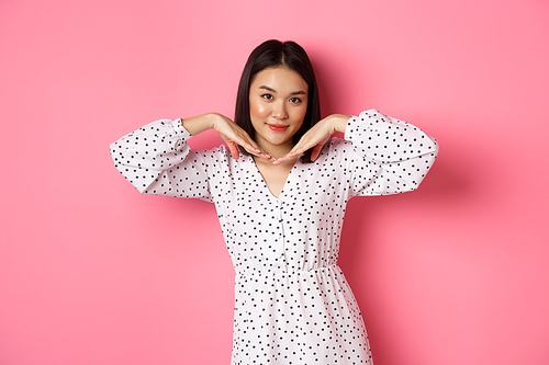 Beautiful asian woman in trendy dress making cute face, holding hands near jawline and gazing coquettish at camera, standing over pink background.