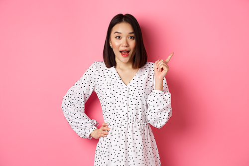 Excited pretty asian girl pointing at upper right corner, gasping amazed and looking at camera, discuss store promo offer, standing over pink background.