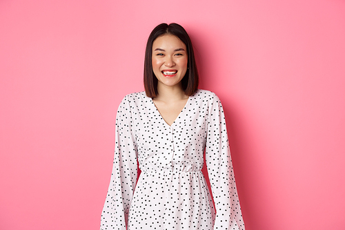 Happy korean woman in dress looking at camera, smiling and laughing with sincere expression, standing over pink background. Copy space