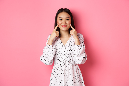 Cute blushing asian girl poking cheeks, smiling happy at camera, wearing romantic white dress, standing over pink background.