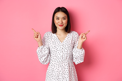 Beautiful asian woman making choice on shopping, pointing fingers sideways and showing variants, smiling at camera, standing over pink background.