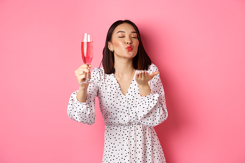 Romantic asian woman raising glass of champagne and sending air kiss at camera, celebrating and having fun, standing over pink background.