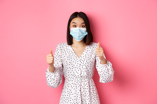 Covid-19, quarantine and lifestyle concept. Excited asian woman in face mask showing thumbs-up, praising good job, looking amazed at camera, standing over pink background.