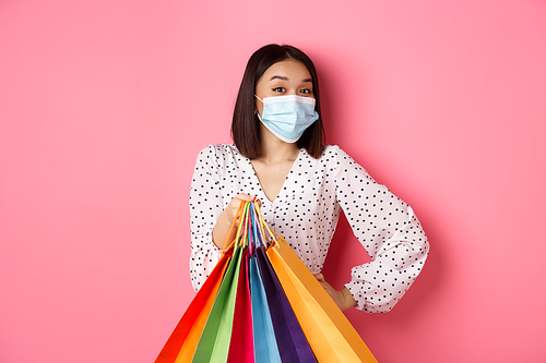 Covid-19, pandemic and lifestyle concept. Beautiful asian woman in medical mask going shopping, holding bags and smiling with eyes, standing over pink background.