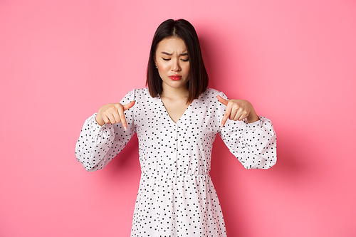 Concerned asian woman peeking down and pointing hand at disturbing banner, frowning upset, standing over pink background.