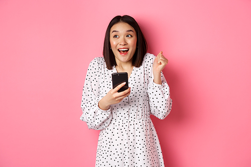 Online shopping and beauty concept. Excited asian woman winning in internet, holding smartphone and rejoicing, smiling happy and celebrating, standing over pink background.