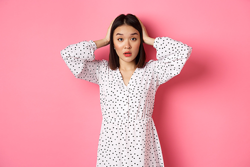 Shocked and troubled asian woman looking confused, holding hands on head, having problem, standing over pink background.