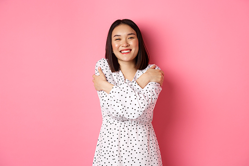 Beautiful asian girl embracing own body, hugging herself and smiling with happy face, looking at camera carefree, standing over pink background.