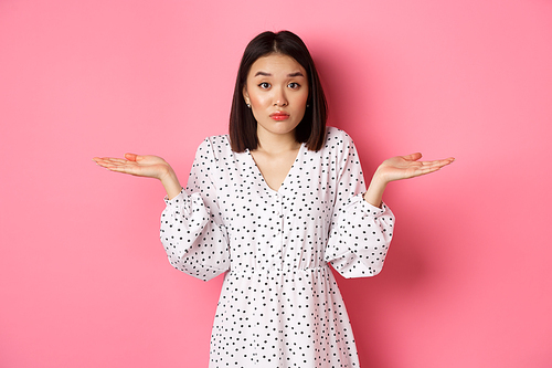 Indecisive cute asian woman shrugging, holding hands spread sideways on copy spaces, dont know, having doubts, standing over pink background.