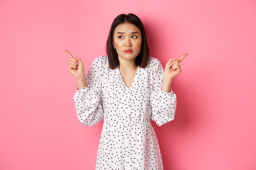 Confused asian woman hesitating, shrugging and pointing hands two sides, staring indecisive and need help with choice, standing over pink background.