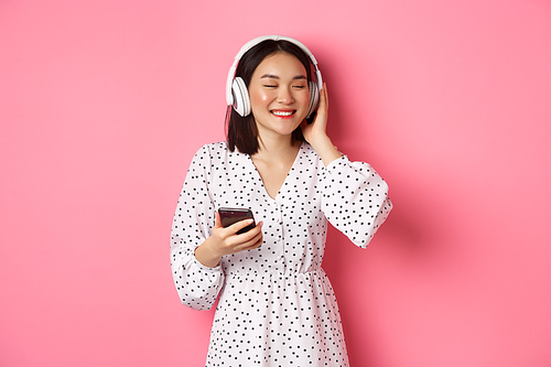 Romantic asian girl listening music in headphones, smiling with closed eyes, holding mobile phone, standing over pink background.