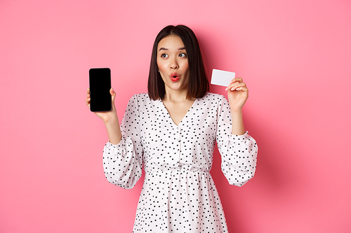 Cute asian woman shopping online, showing bank credit card and mobile screen, smiling and looking left at copy space, standing over pink background.