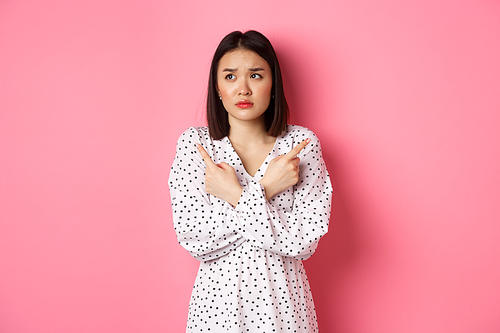 Concerned asian woman having doubts, pointing sideways and looking left with hesitant and sad face, need help with choice, standing over pink background.