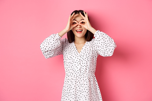 Carefree young asian woman looking through hand binoculars at camera, staring at discounts, standing over pink background.