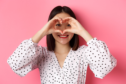 Beauty and lifestyle concept. Close-up of lovely asian woman showing heart sign, smiling and feeling romantic on valentines day, standing over pink background.