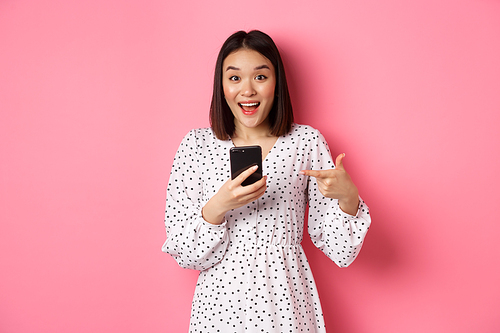 Online shopping and beauty concept. Amazed and happy asian woman pointing at mobile phone, talking about internet promo offer or app, standing over pink background.