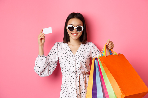 Beautiful asian woman in sunglasses going shopping, holding bags and showing credit card, standing over pink background.