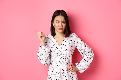 Disappointed asian girlfriend complaining, staring at camera and whining, pointing at upper left corner copy space, standing in dress over pink background.