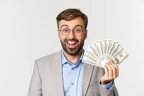 Close-up of happy male entrepreneur in glasses and gray suit, receiving income from business, showing money, standing over white background.