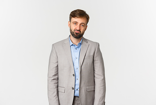 Portrait of skeptical and unamused bearded male employee in grey suit, looking disappointed and sad, standing over white background.