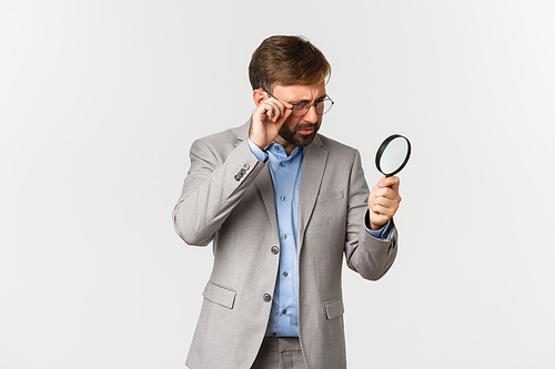 Portrait of serious businessman with beard, wearing grey suit and glasses, searching for something with magnifying glass, investigating over white background.