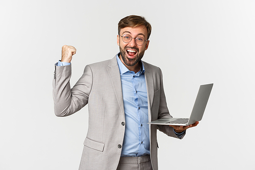 Portrait of happy businessman in suit and glasses, holding laptop and smiling satisfied, saying yes and making fist pump sign, achieve goal at work, standing over white background.