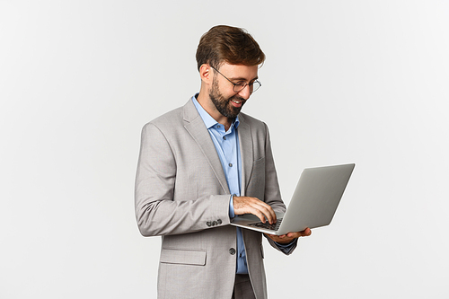 Image of handsome successful businessman working, standing in grey suit and using laptop, standing over white background.