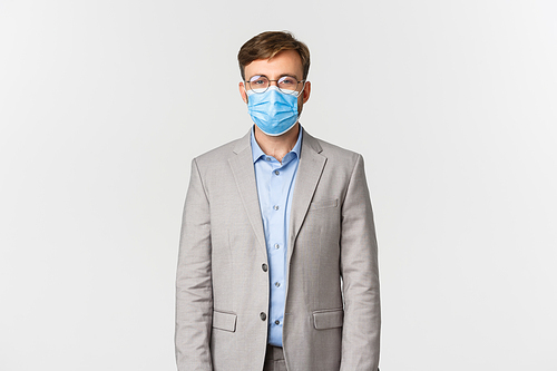 Concept of work, covid-19 and social distancing. Businessman in grey suit and medical mask looking at camera, protect himself from catching coronavirus, standing over white background.