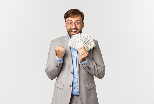 Successful bearded businessman in grey suit and glasses, winning cash, holding money and saying yes, standing happy over white background.