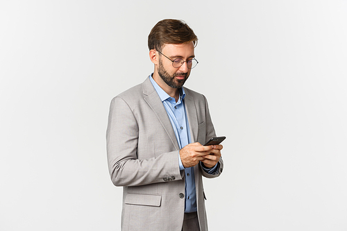 Portrait of handsome businessman with beard, wearing glasses and grey suit, using mobile phone application, writing message, standing over white background.