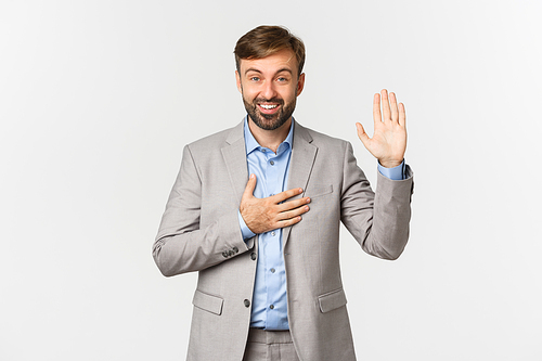 Portrait of smiling bearded businessman making promise, raising hand up and put palm on heart, swearing to tell truth, being honest, standing over white background.