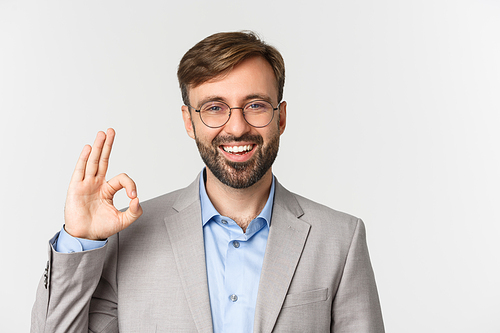 Close-up of handsome bearded man in gray suit and glasses, recommend something, showing okay sign and smiling, give approval, standing over white background.