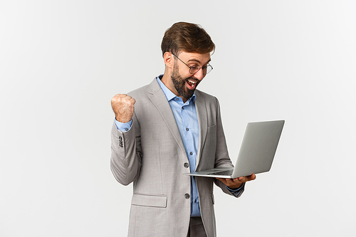 Portrait of happy businessman in suit and glasses, looking at laptop screen and rejoicing, achieving goal, standing over white background.