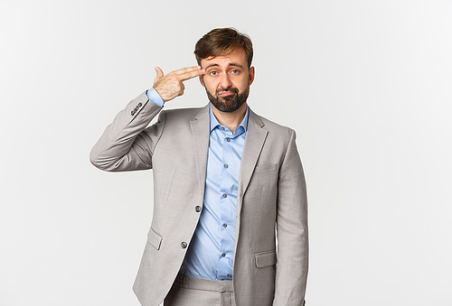 Image of miserable businessman with beard, wearing grey suit, making finger gun sign over head and shooting himself, standing distressed over white background.