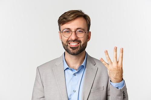 Close-up of handsome bearded man in glasses and gray suit, showing number four and smiling, standing over white background.