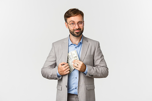 Portrait of confident and successful bearded businessman, wearing grey suit and glasses, putting money in inner pocket, smiling pleased, standing over white background.