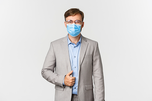 Concept of work, covid-19 and social distancing. Image of confident and successful businessman in gray suit, glasses and medical mask, smiling pleased, standing over white background.