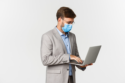 Concept of work, covid-19 and social distancing. Image of working businessman in medical mask and suit, using laptop and standing over white background.