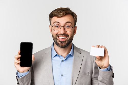 Close-up of handsome smiling male entrepreneur, wearing glasses and gray suit, showing credit card and mobile phone screen, looking excited, standing over white background.