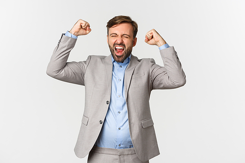 Portrait of successful bearded businessman in grey suit, winning something or achieving goal, shouting for joy and triumphing, celebrating victory over white background.