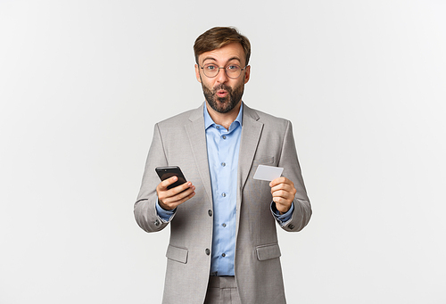 Portrait of excited businessman holding credit card and mobile phone, shopping and paying online, standing over white background.