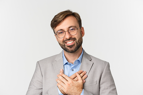 Close-up of handsome businessman in gray suit and glasses, holding hands on heart and thanking for something, smiling grateful, standing over white background.