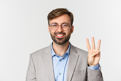 Close-up of handsome bearded man in glasses and gray suit, showing number three and smiling, standing over white background.