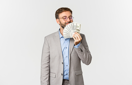 Portrait of successful happy businessman, wearing grey suit and glasses, smelling money and smiling delighted, standing over white background.